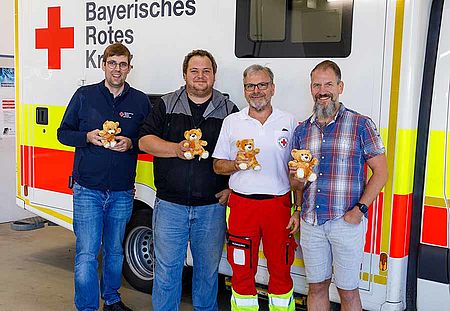 Vor einem Rettungswagen des BRK stehen vier Männer mit kleinen Teddybären in der Hand und fröhlichem Gesichtsausdruck.