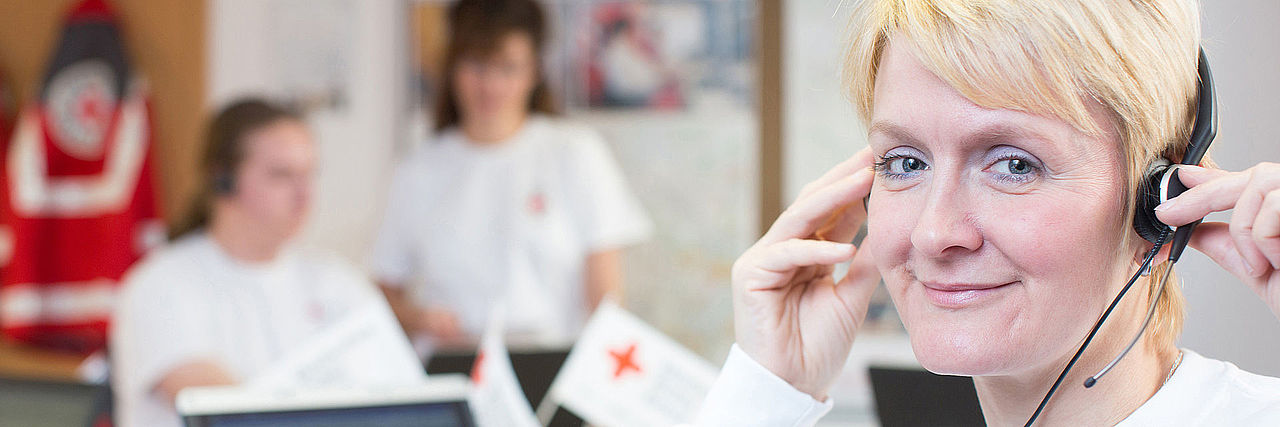 Eine Frau sitzt freundliche lächelnd in einem Servicecenter des DRK / BRK. Sie hat blonde kurze Haare und ein Headset. 