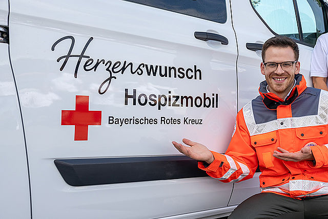 Der Comedian Martin Frank kniet vor dem Herzenswunsch Hospizmobil und präsentiert das Logo an der Seite des Fahrzeugs