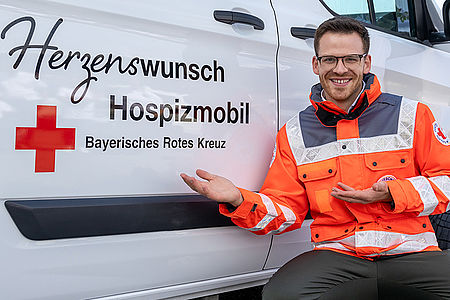 Martin Frank kniet in orangener BRK-Einsatzkleidung neben dem Herzenswunsch Hospizmobil und deutet auf dessen Logo. Er lächelt freundlich.