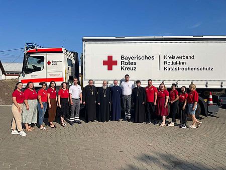 Der große Wechsellader mit dem Logo des BRK steht im Hintergrund. Im Vordergrund Mitglieder des rumänischen Roten Kreuzes und der rumänisch Orthodoxen Kirche und zwei ehrenamtliche Helfer des BRK-Kreisverbandes Rottal-Inn